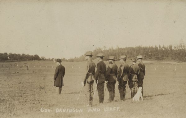 Three-quarter view from rear of group of men standing in a field. Governor James O. Davidson stands in front of the group on the left. Behind him six men in military uniform stand in a line with sabres at their side, with a white dog sitting behind them. Small white markers dot the field in the background. Trees are along a hill in the distance.