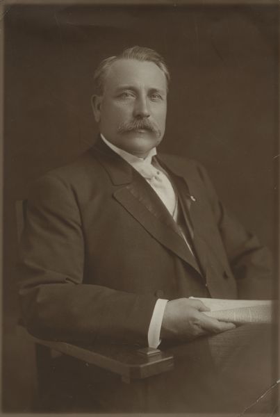 Waist-up portrait of James O. Davidson, sitting. He is wearing a dark suit with a white tie, and is holding a newspaper in his right hand.