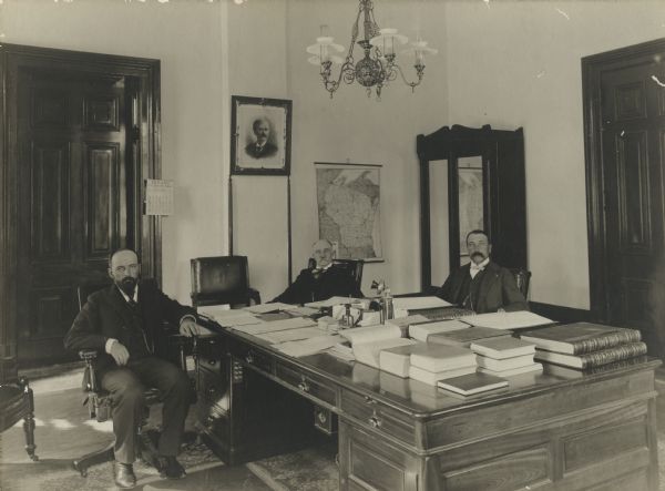Three men are sitting in an office around a large desk with stacks of  books, papers and a telephone. They men are wearing suits, ties and watch fobs, and James O. Davidson is on the far right. Several books and papers lay on the desk in the middle of the room, along with a telephone. There is a map of Wisconsin hanging on wall behind the men.