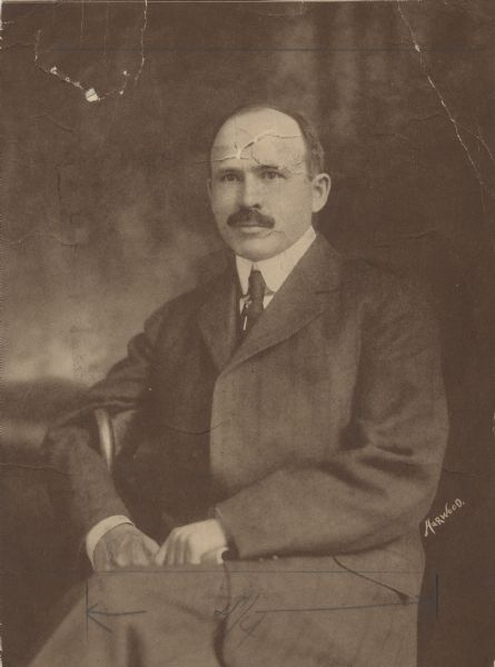 Three-quarter length portrait of Francis E. McGovern sitting slightly sideways in a chair with his right arm hooked around the back. He holds an object in his hands.
