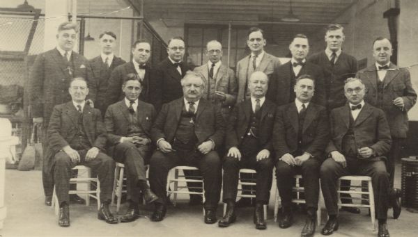 Outdoor group portrait of men from the Mi Lola Cigar Company, along with Emmanuel Philipp, the former governor of Wisconsin (front row third from the left), and his son Cyrus Philipp (second from the left). A number of the men are holding cigars. In the background is a fence and an industrial building.