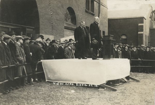 Governor Emmanuel Philipp stands on a dais, covered with a white sheet, behind University of Wisconsin-Madison's President Charles Van Hise. Van Hise stands behind a podium holding his hat in his right hand. A semi-circular iron fence surrounds the dais and crowded behind it stands a crowd of young men wearing coats, hats (some are flat caps, some are brimmed), and ties. The group is in front of a brick building with arched doorways and windows. A sign on the building reads: "Armory and Gymnasium of the University of Wisconsin." There is another brick building in the background on the right.