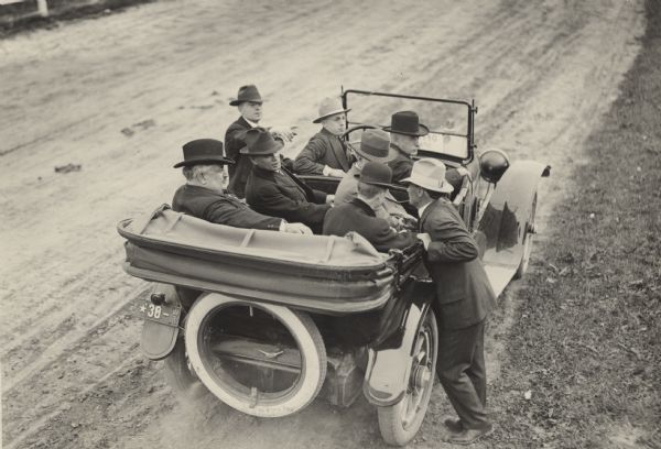 Slightly elevated view from rear of Governor Philipp sitting in a Pierce Arrow luxury car in the back seat on the left, with the top down. Five other men sit in the car with him, and two stand next to it. All of the men are turned towards the man leaning against the car on the right. They are stopped along the side of an unpaved road.