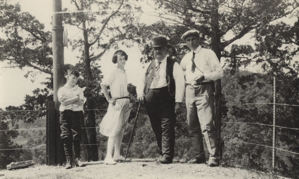 Group portrait of four people standing in front of a wire fence on the edge of a cliff, with trees and a valley in the background. Walter Alexander stands furthest on the far right, next to Governor Emmanuel Philipp. Alexander wears a shirt and tie with a flat cap. Philipp holds a cane and wears a shirt, tie and vest with a brimmed hat. A young girl stands next to Philipp and is wearing a knee-length dress and stockings. On the far left a young boy is leaning against a post with his arms crossed.