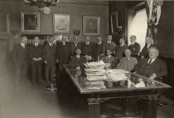 Governor Emmanuel Philipp sits at his desk in a group portrait with his senators. Three other men sit in chairs behind the desk, and eleven men stand in a row behind them. Papers and telephones sit on the glass topped desk. On the left is a spittoon on the floor near the desk. Along the rear wall are bookshelves, and framed paintings and a clock are hanging on the wall. A flag hangs above Philipp's head next to a window flanked by wall sconces.