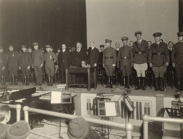 View from audience towards stage where Governor Emmanuel Philipp is standing behind a desk. Standing with him, in front of their chairs, are two men in suits, as well as rows of men dressed in military clothes. In front of the stage is a piano, and four stands with sheet music displayed.