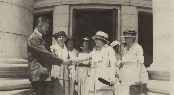 Governor John J. Blaine (left) shaking the hand of Mabel Raef Putnam surrounded by other women of who have come to see the governor sign the Women's Rights Bill. Ms. Putnam is holding a quill pen in her left hand. The pen was supplied by the National Women's Party to be used in signing the Bill. They are standing in front of an entrance to the Wisconsin State Capitol. From left to right: Governor Blaine, Mrs. Max Rotter, Rachel Jastrow, Mrs. Kathryn Hoebel, Mabel Raef Putnam, unidentified, unidentified (possibly Mrs. Anna Blaine), Mrs. Louise Anderson.