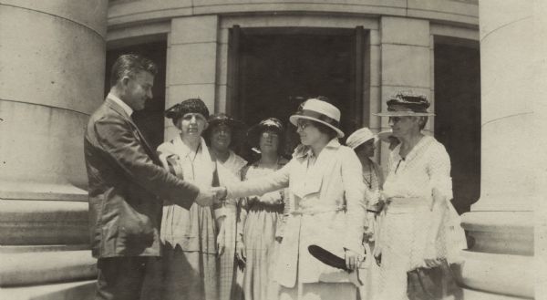 Governor John J. Blaine (left) shaking the hand of Mabel Raef Putnam surrounded by other women of who have come to see the governor sign the Women's Rights Bill. Ms. Putnam is holding a quill pen in her left hand. The pen was supplied by the National Women's Party to be used in signing the Bill. They are standing in front of an entrance to the Wisconsin State Capitol.
From left to right: Governor Blaine, Mrs. Max Rotter, Rachel Jastrow, Mrs. Kathryn Hoebel, Mabel Raef Putnam, unidentified, unidentified (possibly Mrs. Anna Blaine), Mrs. Louise Anderson.