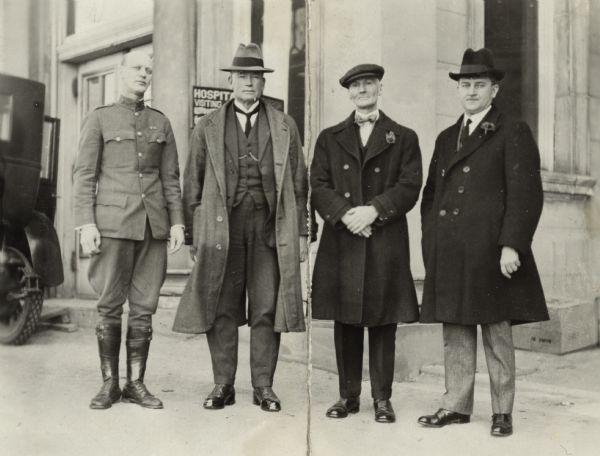 Group portrait of men standing outside of a hospital entrance. From right to left: John J. Blaine, Wisconsin's 24th governor; Albert A. Fuller of Albert A. Fuller & Sons Stock Farm, North Lake, Wis; David C. Pryce, Gen. Farms Mgr., Nestle Anglo-Swiss Condensed Milk Co. of Austral-Asia; Thomas J. Heldt, Clinical Dir., U.S. Pub. Health Service Hospital No. 37, Waukesha, Wis. Blain, Fuller, and Pryce wear suits, coats and hats, while Heldt wears a military uniform without a hat.  They stand in front of the stone building; a sign behind Pryce reads "Hospital."