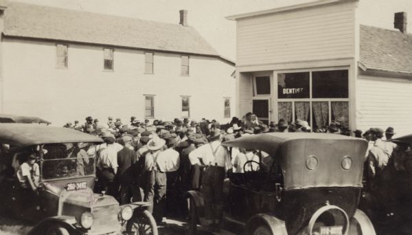 Slightly elevated view towards a crowd of men gathered in front of a dentist building. John J. Blaine, Wisconsin's 24th governor stands speaking near the entrance of the small storefront. Automobiles are in the foreground, and a large, two-story wood-sided building is in the background on the left.