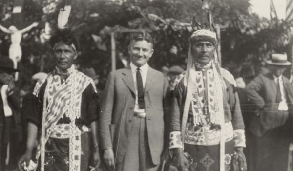 Outdoor group portrait of John J. Blaine between two men in Chippewa clothing and headdresses. Other men in suits and hats stand or walk in the background. There is a large crucifix behind them on the left.