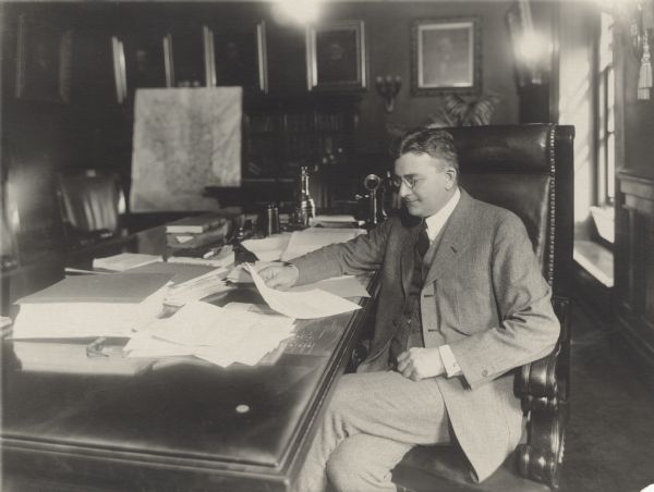 Governor John J. Blaine, Wisconsin's 24th governor, sits in an office next to a desk in a leather chair. Papers, books and a telephone are on the desk. On the back wall is a bookcase, and hanging on the wall are framed portraits of men. A map of Wisconsin sits on a stand near the back corner of the room.