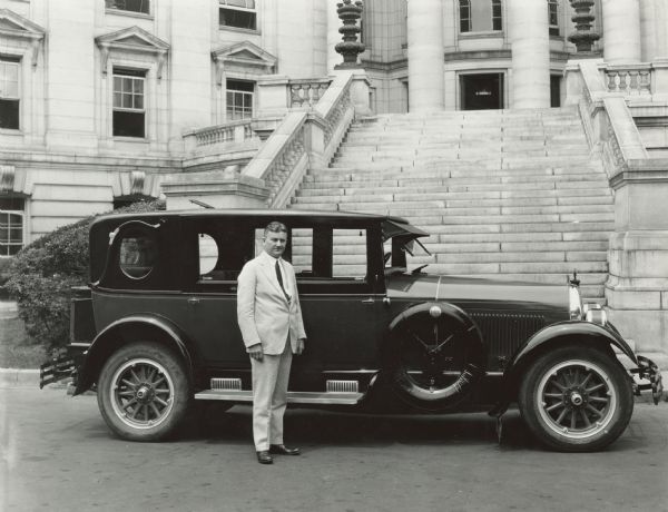 John J. Blaine is standing outside the entrance steps to the Wisconsin State Capitol building in front of a Kissel automobile. He is wearing a light-colored suit. The car is black, with the companies brand on the spare tire to Blaine's right.