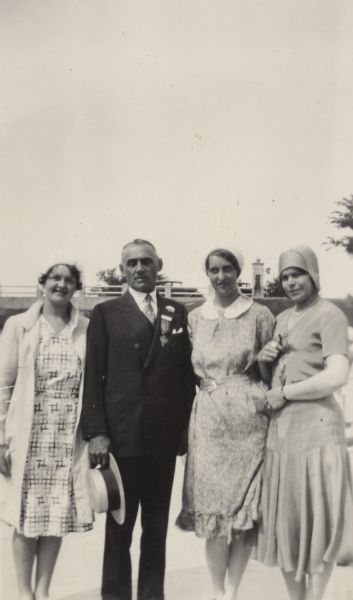 Governor Walter Kohler, Sr. stands for a portrait with three women at the National American Legion Convention. From right to left: Marcella Bartlein, Bernice Weigle, Governor Kohler, Jacquline Meyers. The women wear knee length dresses and caps and Meyers wears a coat. A medal is pinned on Kohler's left lapel with a small flower above it. Bartlein holds a similar medal in her right hand. Behind the group are trees and a bridge.