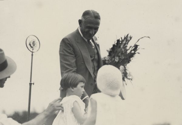 Walter Kohler Sr. holding a bouquet under his left arm as he looks down at two young girls in front of him. Both girls wear sleeveless dresses. A man wearing a hat is on the far left and is extending a hand to touch the back of the girl on the left. Behind Kohler is a tall ring microphone (also called spring microphones or double button carbon ring microphones).