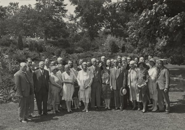Governor Walter J. Kohler Sr. stands for a group portrait with several men and women on a lawn with trees and foliage. He is the fifth person from the right in the first row, wearing a suit with a flower pin on his lapel and holding his hat in his left hand. The women in the group are all in the first or second row and wear knee or mid-calf length dresses and caps or hats. The men are in suits and ties.