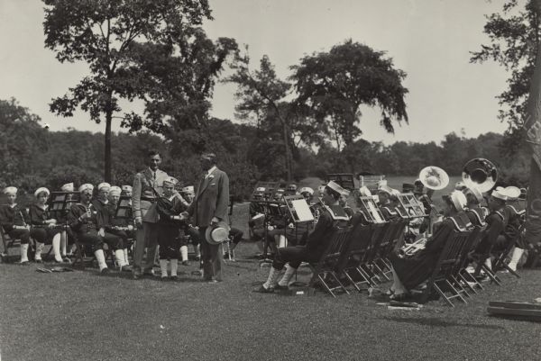 Governor Walter Kohler Sr. shakes the hand of a young boy, Henry Stoehr. They stand next to Henry Winsauer, and are surrounded by members of the Kohler Junior Band. The boys and girls sit with their instruments and sheet music wearing matching uniforms. They are outdoors on a lawn surrounded by trees. Kohler holds his hat in his hand, and is wearing a suit with a flower pin on the lapel.