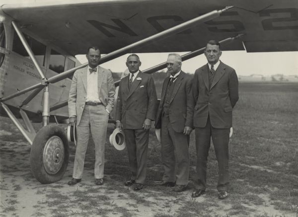 Group portrait of four men standing outdoors under the wing of an airplane. From left to right: F.S. Brandenburg, Governor Kohler, George Charlesworth, and Reverend Adolph J. Soldan. They are all wearing suits and hold hats in their hands.