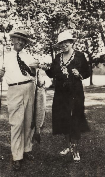 Albert and Katherine Schmedeman are standing outdoors, both holding a small stringer with a large fish hanging from it. Both Albert and Katherine are holding fishing poles in their other hands. Albert is wearing a hat, white shirt, light-colored pants and a polka dot tie. Katherine is wearing a dark-colored mid-calf length dress, hat, and two-tone shoes. In the background are trees along a shoreline.