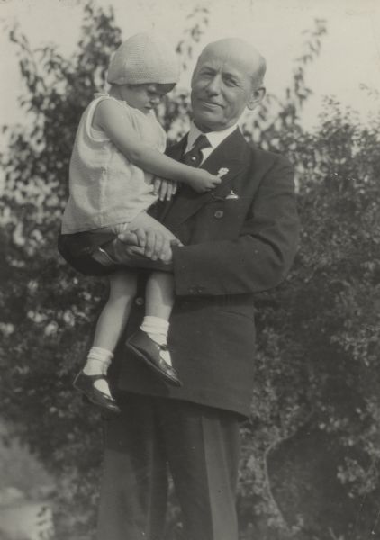 Albert Schmedeman, mayor of Madison (1926-1932), holds a little girl in his arms as they stand outdoors. The girl (identified as Lari Ballam) wears a dress and a cap, and is reaching out to touch a flower pin on Schmedeman's lapel. Schmedeman wears a suit and tie, with a pocket watch in his breast pocket.