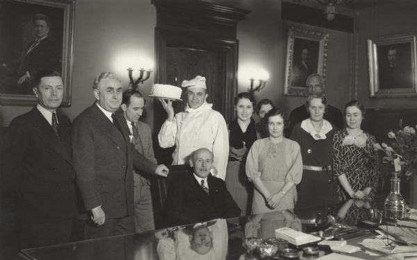 Governor Albert Schmedeman sits at his desk for a group portrait. To his right stand three men wearing suits, and to Schmedeman's left stand five women wearing dresses, with one man standing behind them. Behind the governor a man dressed as a chef holds up a white cake in one hand.