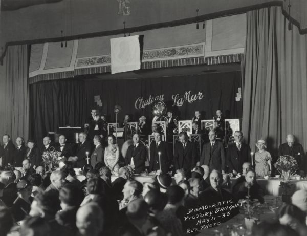 View from audience, sitting at tables in the foreground, towards a group of men and women standing at a long table adorned with flowers and candles. Behind them a brass band plays on a stage. The newly elected Governor Albert Schmedeman stands four people in from the left, next to his wife, Katherine Schmedeman.