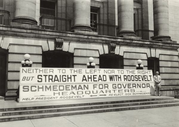 Governor Albert G. Schmedeman stands next to a large campaign sign for his re-election as governor outdoors at the Wisconsin State Capitol. The sign reads: "Neither to the left nor to the right but straight ahead with Roosevelt. Schmedeman for Governor Headquarters. Help President Roosevelt. Re-elect Gov. Schmedeman."