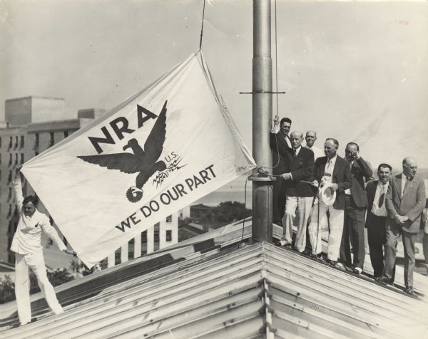 Albert G. Schmedeman stands on a roof of a building with seven other men. He is next to a pole holding the rope that will raise the flag on display. One man stands on the left side of the roof holding the flag taut. The flag reads: "NRA We Do Our Part" (National Recovery Administration).