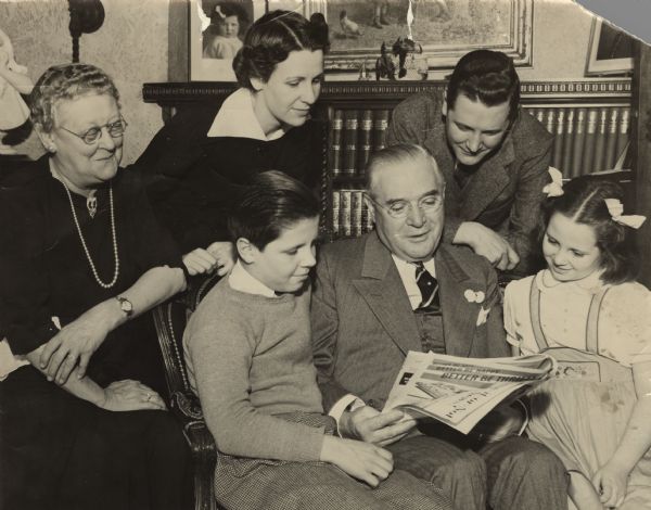 Julius P. Heil sits reading a newspaper with members of his family. To his right is Joe Jr., 13 and to his left is Barbara Ann, 9. His wife, Elizabeth, sits on the left. Next to her stands Elizabeth and Julius' daughter-in-law, Marjorie (née Nichols). Joseph F. Heil, Elizabeth and Julius' son, leans over the chair between Julius Heil and Barbara Ann. In the background is a bookshelf and above are framed pictures and paintings.