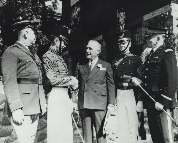 Left to right: Brigadier General Ralph M. Immell; Cadet Lieutenant Howard B. Woodside; Governor Julius Heil; Cadet Captain Henry Fromm; Col. Roy Farrand, head of St. John's Military Academy. Heil is wearing a suit while holding his brimmed hat in his left hand while shaking the hand of Cadet Lt. Howard Woodside. The other four men wear military uniforms and the cadets wear a shako (a plumed military hat). Col. Roy Farrand holds a cane.