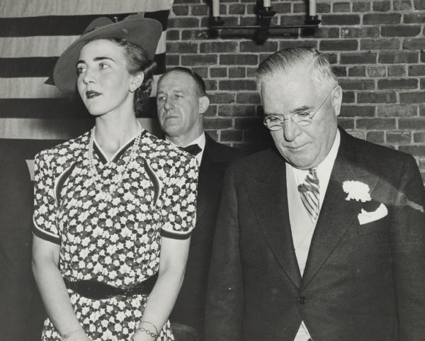 Princess Ingrid of Denmark with Wisconsin Governor Julius Heil. The princess is wearing a floral patterned short sleeved dress, a pearl necklace, earrings, bracelets, a small jeweled watch, and a hat. The governor is wearing a suit and tie with a flower pin on his lapel. A man in a suit and bow tie stands behind them. There is an American Flag on the brick wall in the background.