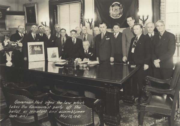 Governor Julius P. Heil sits at his desk signing a piece of paper. Behind him stand senators and assemblymen. A vase of flowers sits on the left corner of the desk, a clock, desk calender and small glass bowls sit in the middle. The Wisconsin state flag hangs on the wall behind the desk, and the American flag is on the left. Written on the image is: "Governor Heil signs the law which takes the Communist party off the ballot as senators and assemblymen look on!"