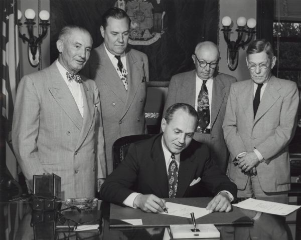 Governor Walter J. Kohler, Jr., sits at his desk signing a document. Four men stand behind him, all wearing suits. The Wisconsin state flag hangs on the wall in the background. The American flag hangs from a pole along the far left.