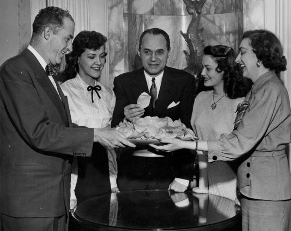 Governor Walter J. Kohler Jr. stands behind a small round table holding a (tortilla?) chip in his right hand and bracing his left fist on the table. Two women stand to his left, and a woman and a man stand to his right, all holding the platter of tortilla chips with a bowl of dip. Behind the group is a marble wall with part of a koi statue visible behind the governor.