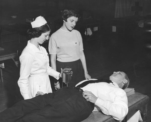 Walter Kohler Jr. lying on a cot, while two women, one in a nurse's uniform, stand beside him on the left. The nurse holds a glass bottle used to collect blood. More cots and medical prep tables are visible in the background. The Red Cross flag hangs in the upper right corner.