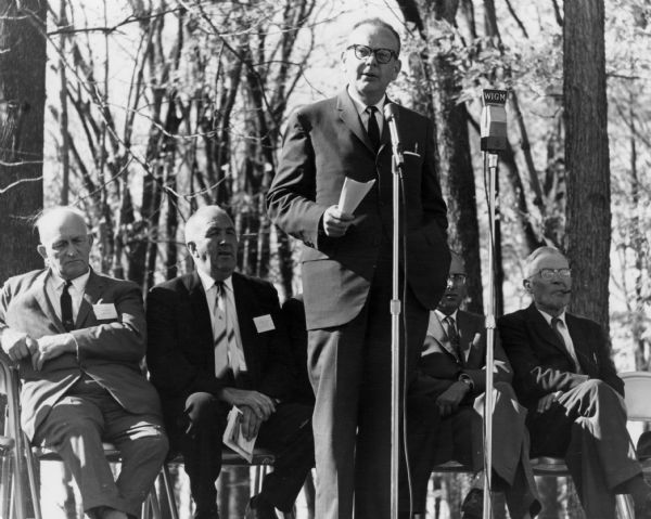 Governor John W. Reynolds stands on a stage in front of two microphones, one labeled WIGM. He holds a piece of paper in his right hand. Four men sit behind him. In the background are trees.