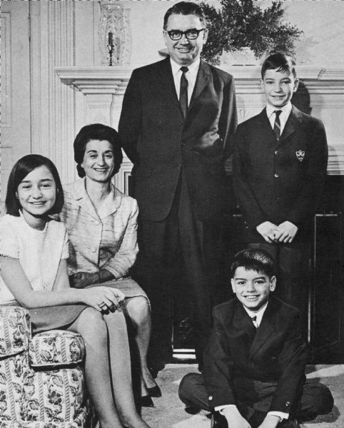 Lieutenant governor Patrick J. Lucey stands in front of a fireplace with his hands behind his back. To the right are his two sons, Paul and David. To the left, Jean Lucey (wife) and Laurie Lucey (daughter) sit on a couch. The boys wear suits, Mrs. Lucey wears a suit dress, and Laurie wears a blouse and skirt.