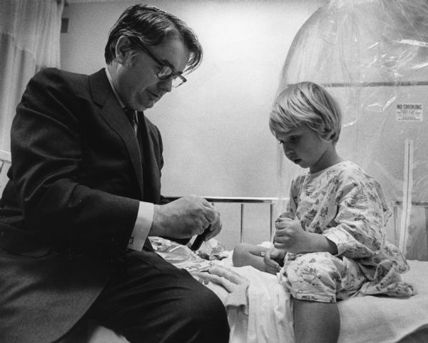 Governor Patrick J. Lucey sitting on a hospital bed holding something in his hands. A young boy, Douglas Huete, is sitting on the bed wearing a hospital gown and looking at the object in the governor's hands. Behind the boy is a plastic sheet or bubble. A sign reads: "No Smoking."