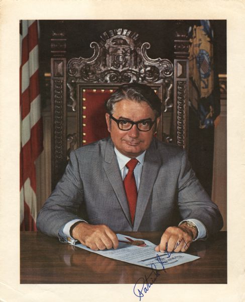 Signed portrait of Governor Patrick J. Lucey, Wisconsin's 38th governor. He is sitting in an elaborate chair behind a desk. In the background hangs the Wisconsin Flag and American Flag. Lucey rests his arms on desk over a document bearing a seal. The photograph is autographed by Governor Lucey.