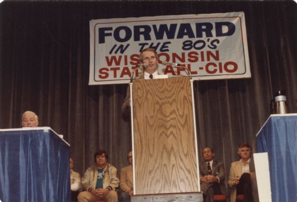 Governor Anthony S. Earl stands at a podium on a stage. A man sits at a table draped in blue cloth on the left. Behind Earl sit several men in chairs. Some of the men are wearing suits, but one man is wearing a gold jacket, a t-shirt that reads "Earl" and tan pants. A banner on the curtain in the background reads: "Forward in the 80's. Wisconsin State AFL-CIO." (American Federation of Labor - Congress of Industrial Organizations).