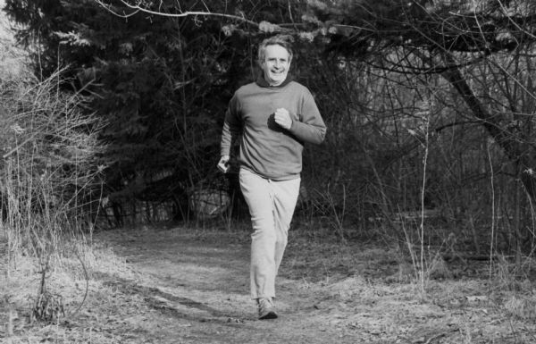Governor Anthony Earl jogging through a dirt path in a wooded area. He is wearing a sweatshirt, pants and sneakers.
