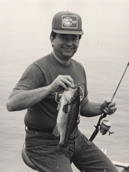 A young Governor Thompson sits on a chair in a boat holding a bass in his right hand and a fishing pole in his left. He wears a t-shirt, jeans, and a baseball cap that reads "Wisconsin Fish Management = Fantastic Fishing."