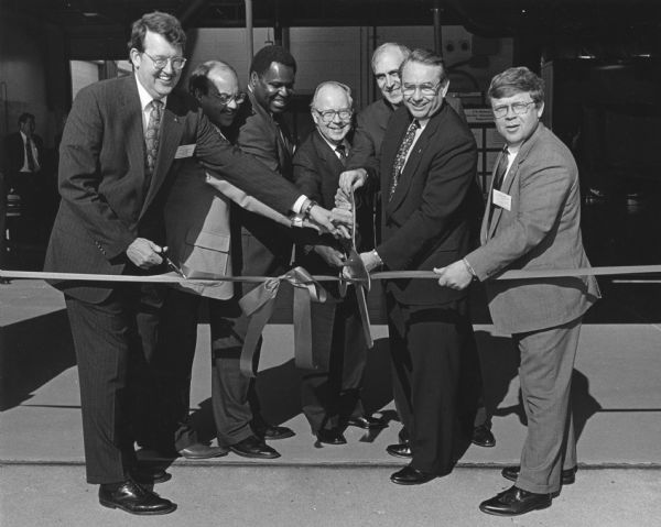 Governor Tommy Thompson (second from the right) holds a large pair of scissors ready to cut a long ribbon. Six other men stand in with him, all (except for the man furthest to the right) holding the large scissors. The man furthest to the left holds a pair of regular sized scissors. A sign behind the governor, partially obscured, reads: "UW-Milwa_____ __nter for Alternat_."