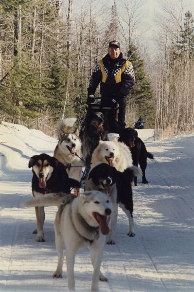 Governor Tommy Thompson driving a sled pulled by seven dogs of various breeds, at least two of which are huskies. Snow covers the ground and trees line the path. A person on a snowmobile is in the far background.