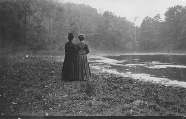 Two women are standing with linked arms with their backs to the camera. They are standing on the grass and dirt shore of a lake surrounded by forest.