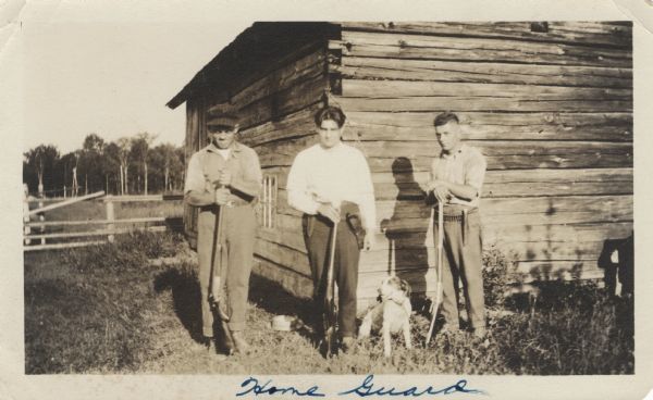 Three men posing standing in front of a log house belonging to the Getto family, all holding guns. A dog is sitting on the ground between two of the men. The man on the far left is wearing a plaid cap, and the other two men are wearing shirts and pants with knives or guns attached to their belts.