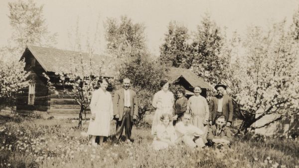 Members of the Getto family posing in front of a log house surrounded by flowering trees. The three people sitting in the front row are, left to right: Maria Getto, Henry Ketola, and Henry Getto. The six people standing in the back row are, left to right: Senia Getto Aho, Herman Aho, Lydia Hovi, Ruth Howland, Alma Getto, and Oscar Getto.