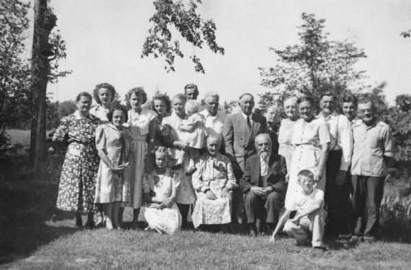 Group portrait of men, women, and children posing outdoors. The men are wearing suits and the women are wearing dresses (mostly short sleeved and mid-calf length). Trees are in the background. On the left is a dinner bell on the top of a pole, and behind it is a birdhouse on a tree.