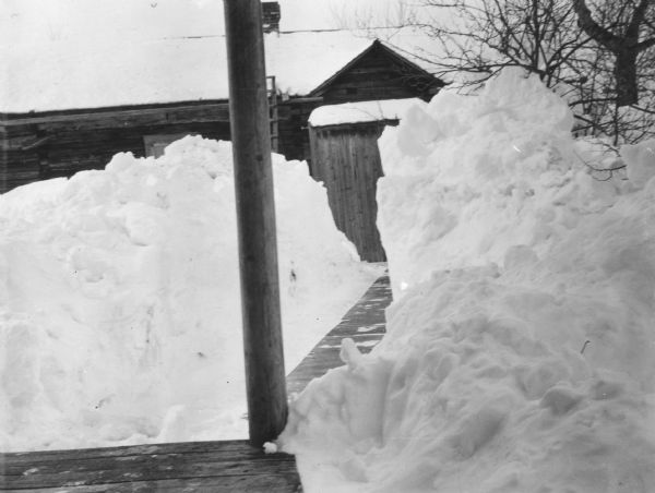 View from porch of a wood sided building behind a large pile of snow. A path has been shoveled on the wood walkway between the porch and the outer building.