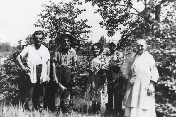 Outdoor group portrait of five members of the Getto family posing among trees. Left to right: Fred Getto, Henry Getto, Alma Getto, Oscar Getto, and Maria Getto. The women wear dresses, and the men wear shirts and pants. Henry Getto also wears suspenders and is holding a newspaper in his right hand. A log house is in the background.